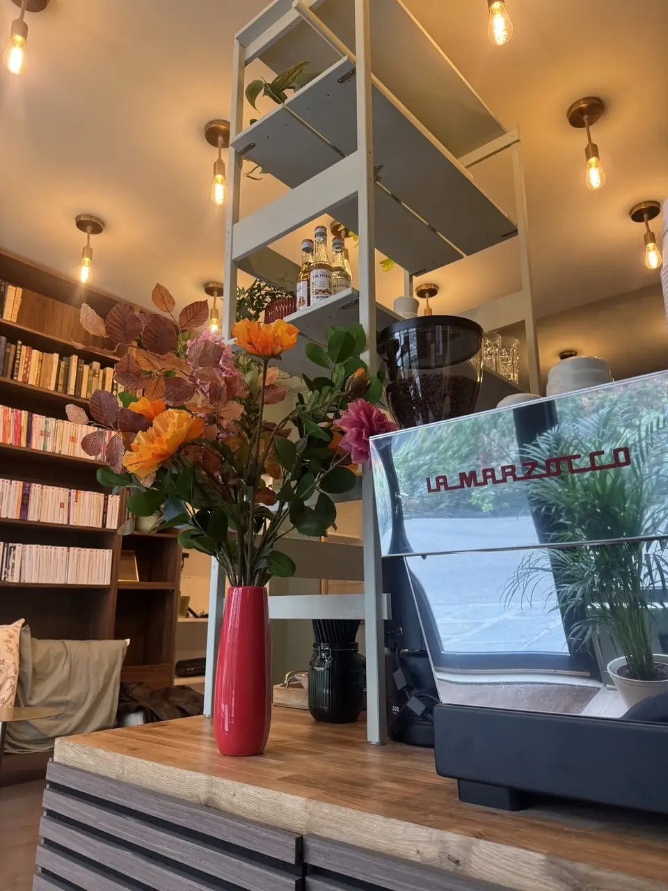 Le comptoir en bois clair avec la machine à espresso La Marzocco, un moulin à café et un bouquet de fleurs dans un vase rouge